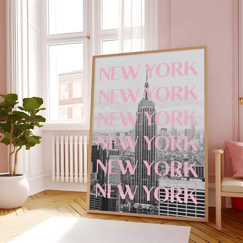 May include: A framed print featuring a black and white photograph of the New York City skyline, with the Empire State Building prominently displayed. The word "NEW YORK" is repeated in pink over the image. The frame is a light wood color.