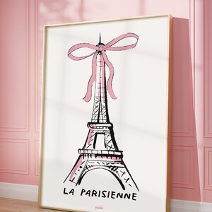 May include: A black and white illustration of the Eiffel Tower with a pink bow on top. The text "LA PARISIENNE" is below the tower. The image is framed in gold.