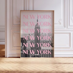 May include: A framed black and white photograph of the New York City skyline, featuring the Empire State Building. The word "NEW YORK" is repeated in pink over the image. The frame is gold.