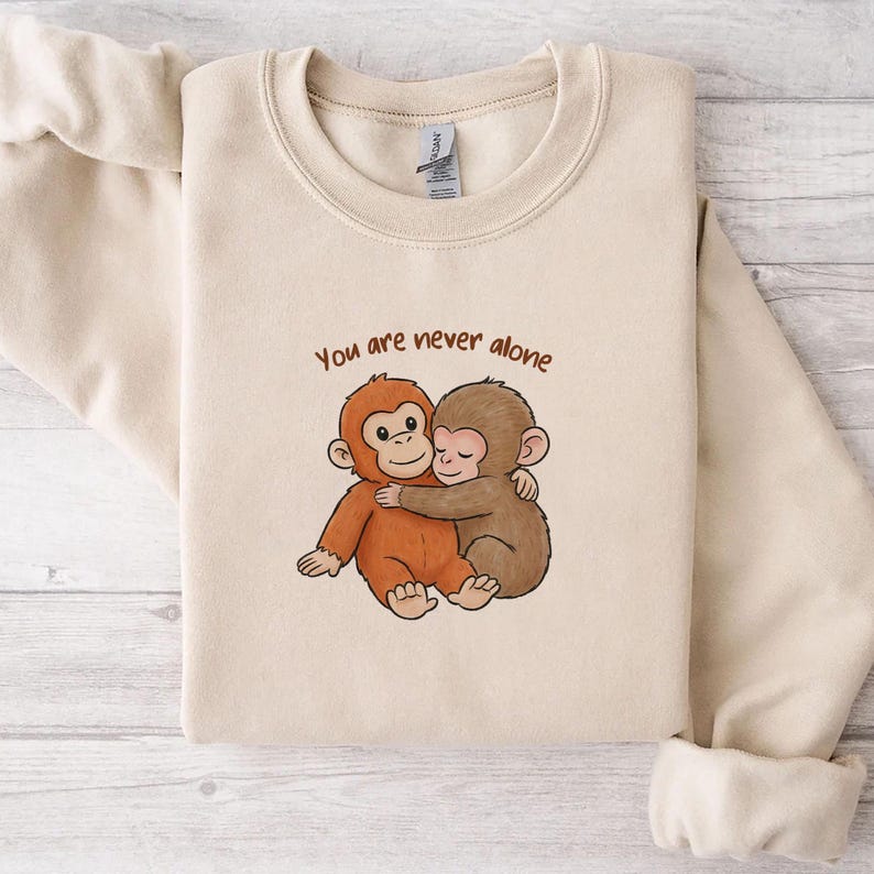 May include: A beige sweatshirt featuring a cartoon illustration of two hugging monkeys. The text "You are never alone" is above the monkeys in brown lettering. The sweatshirt is made of a soft material.