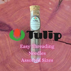 Puede incluir: Un tubo de ensayo de vidrio transparente con tapón de corcho que contiene agujas de metal surtidas de fácil enhebrado. La etiqueta del tubo muestra el logotipo de la marca Tulip y el texto "Easy-Threading Needles Assorted".