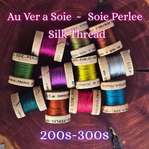 Puede incluir: Una colección de coloridas bobinas de hilo de seda, cada una con una base de madera y etiquetas de texto. Las bobinas muestran varios tonos, incluyendo verde, morado y naranja. El texto "Au Ver a Soie ~ Soie Perlee Silk Thread" es visible.