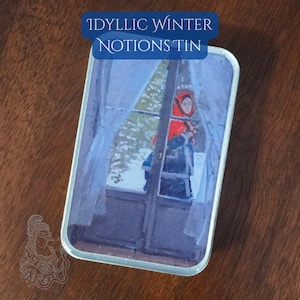 May include: A rectangular metal tin with a lid featuring a painting of a person in a red hat looking out a window at a snowy scene. The text "Idyllic Winter Notions Tin" is displayed on the lid in a blue banner.