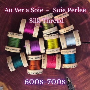 May include: A collection of colorful silk thread spools, each with a wooden base and labeled with text. The spools display various shades, including green, purple, and gold. The text "Au Ver a Soie ~ Soie Perlee Silk Thread" and "600s-700s" is visible.