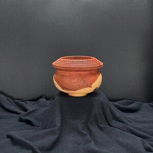 May include: A wooden bowl with a reddish-brown color and a natural wood grain pattern. The bowl has a rounded shape and a slightly flared rim. It is sitting on a black fabric surface.