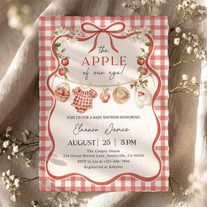 May include: A rectangular baby shower invitation with a red and white gingham border. The center features a clothesline with a basket of apples, a gingham romper, an apple, a straw hat, and a white onesie. Text reads: The APPLE of our eye!