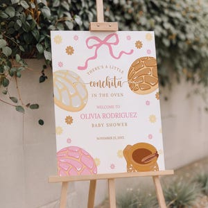 May include: A white rectangular baby shower sign on a wooden easel featuring a pink ribbon, yellow flowers, and illustrations of concha sweet breads in blue, brown, and pink. Text reads: There's a little conchita in the oven, Welcome to Olivia Rodriguez Baby Shower, November 23, 20XX.