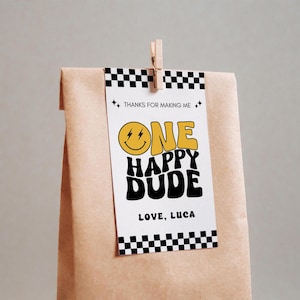 May include: A gift tag with the text "ONE HAPPY DUDE" in yellow and black, with a smiley face. The tag is attached to a brown paper bag with a wooden clip. The tag also says "THANKS FOR MAKING ME" and "LOVE, LUCA".