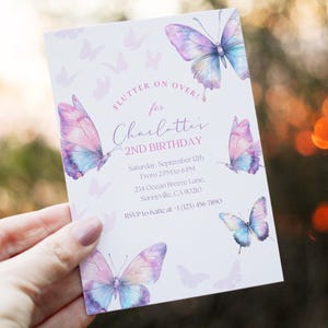 Peut inclure: Une invitation rectangulaire blanche ornée de papillons aquarelles violets, roses et bleus. Le texte "FLUTTER ON OVER! for Charlotte's 2ND BIRTHDAY" est visible. La date, l'heure et le lieu sont également indiqués.