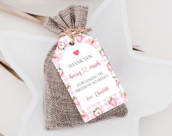 Puede incluir: Una pequeña bolsa de regalo de arpillera atada con hilo, con una etiqueta con un patrón de cuadros rosa y ilustraciones de fresas. La etiqueta dice "Thank You berry much" y "For making my birthday so sweet."