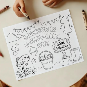 以下が含まれることがあります： 「Jackson is O-Fish-Ally One」のテキストが入った白黒の塗り絵。魚、魚の入ったバケツ、釣り看板、数字の1の風船、その他の釣りテーマのイラストが描かれています。クレヨンが見えます。