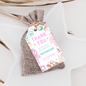 May include: A small burlap bag tied with twine, holding a gift. A white tag with watercolor illustrations and text that reads "Thank You" and "For making my birthday SPATACULARI! Love, Madison."