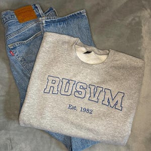 May include: A grey sweatshirt with the word "RUSVM" in blue lettering and the text "Est. 1982" is laid over a pair of light blue denim jeans. The jeans have a brown leather tag.
