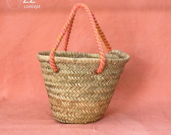 May include: A small, woven straw basket with a pink and orange rope handle. The basket is sitting on a peach-colored surface.