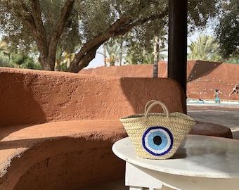 May include: A woven straw tote bag with a blue and white evil eye design. The bag is sitting on a white table with a brown, curved bench in the background.