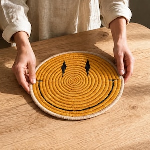 May include: A round, coiled fiber mat featuring a yellow smiley face design. The mat is constructed with concentric circles of yellow material, accented by two black stitched diamond-shaped eyes and a wide, curved black stitched mouth.