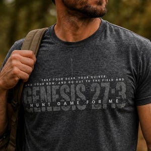 May include: Dark gray t-shirt with the text "GENESIS 27:3" in a distressed font. The shirt also features the quote "TAKE YOUR GEAR, YOUR QUIVER, AND YOUR BOW, AND GO OUT TO THE FIELD AND HUNT GAME FOR ME."