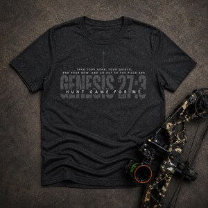 May include: Dark gray t-shirt with the text "TAKE YOUR GEAR, YOUR QUIVER, AND YOUR BOW, AND GO OUT TO THE FIELD AND GENESIS 27:3 HUNT GAME FOR ME." A camouflage bow is positioned to the right of the shirt.