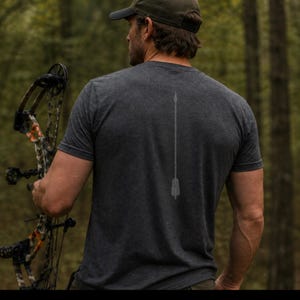 May include: Dark gray t-shirt with a faded arrow design on the back. The person in the image is holding a bow and arrow. The background is a blurred forest scene.