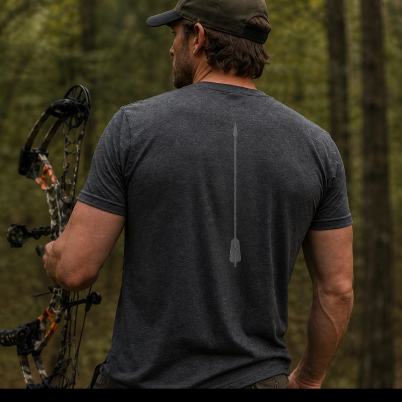 May include: Dark gray t-shirt with a faded arrow design on the back. The person in the image is holding a bow and arrow. The background is a blurred forest scene.