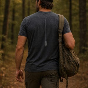 May include: Dark gray t-shirt with a faded spear graphic on the back. The shirt is worn by a person walking in a forest, carrying a green backpack and wearing olive green cargo pants. The shirt's design is simple and minimalist.