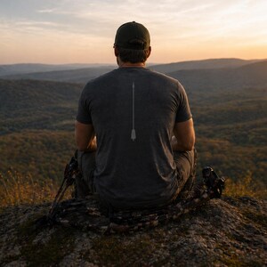May include: A dark gray t-shirt with a white arrow design on the back. The person is wearing a green cap and sitting on a rock, with a bow and arrow set nearby. The background features a scenic view of mountains and a sunset.