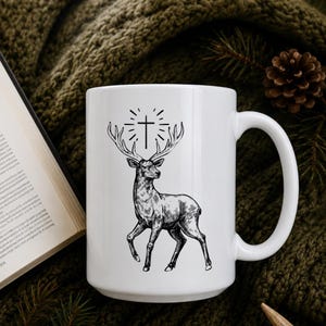May include: White ceramic mug featuring a black and white illustration of a deer with antlers and a cross above its head. The mug has a curved handle and is set against a textured green background.