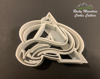 May include: A gray plastic cookie cutter in the shape of a stylized letter "A" with a mountain and text "Rocky Mountain Cookie Cutters". The cutter has multiple layers and a complex design, suitable for baking.
