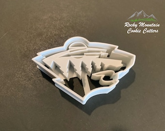 May include: A white plastic cookie cutter shaped like the logo of the NHL's Arizona Coyotes. The logo features a stylized coyote head with a mountain range in the background. The cookie cutter is made by Rocky Mountain Cookie Cutters.