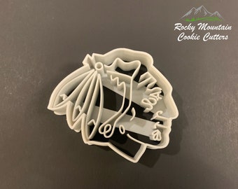 May include: White plastic cookie cutter shaped like a Native American chief wearing a headdress. The cookie cutter is designed to cut out a cookie in the shape of a Native American chief.
