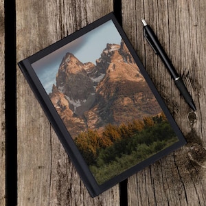 May include: A black journal with a landscape image of a mountain range and forest. A black pen rests on the wooden surface next to the journal. The journal's cover is black, and the image features brown and green tones.