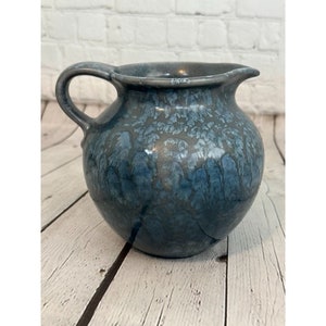 May include: A handcrafted, blue ceramic pitcher with a handle and a spout. The pitcher has a textured, glazed surface with shades of blue and gray. The pottery is likely used for serving beverages.