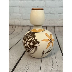 Puede incluir: Un jarrón de cerámica blanco con un diseño geométrico marrón y amarillo y una flor amarilla. El jarrón tiene una base ancha y un cuello estrecho.