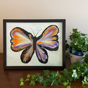 May include: Framed painting of a butterfly with orange, purple, and white abstract paint on a white background.