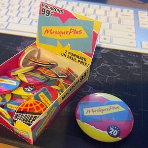 May include: A box filled with colorful round buttons featuring the logo "Musique Plus" and the text "2 formats un seul prix!"  The buttons are red, yellow, blue, and white.  A single button is displayed outside the box.  The button is yellow, red, and blue with the text "Musique Plus" and "20 ans".