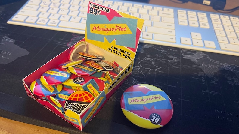 May include: A box filled with colorful round buttons featuring the logo "Musique Plus" and the text "2 formats un seul prix!"  The buttons are red, yellow, blue, and white.  A single button is displayed outside the box.  The button is yellow, red, and blue with the text "Musique Plus" and "20 ans".