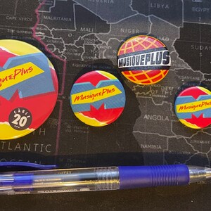 May include: Four round, colorful buttons with the text "Musique Plus" on them. The buttons are arranged on a black surface with a world map in the background. The largest button has the text "C&eacute;l&eacute;br&eacute; 20" on it.