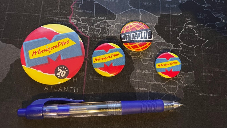 May include: Four round, colorful buttons with the text "Musique Plus" on them. The buttons are arranged on a black surface with a world map in the background. The largest button has the text "C&eacute;l&eacute;br&eacute; 20" on it.