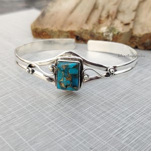 Könnte beinhalten: Ein silbernes Armreif-Armband mit einem Türkis-Stein, der in einen silbernen Rahmen gefasst ist. Das Armband hat ein zartes, verziertes Design.