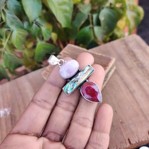 Könnte beinhalten: Ein silberner Anhänger mit drei Edelsteinen. Der obere Edelstein ist rosa, der mittlere Edelstein ist eine grüne und braune Abalone-Schale, und der untere Edelstein ist ein roter Rubin.