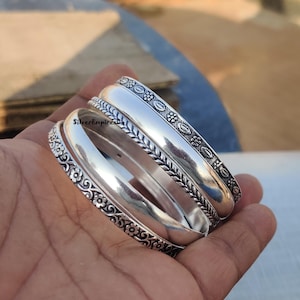 Könnte beinhalten: Drei silberne Armreifen mit aufwendigen floralen und geometrischen Mustern. Die Armreifen sind übereinander gestapelt und haben eine polierte Oberfläche.