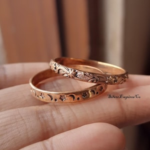 Könnte beinhalten: Zwei goldfarbene Ringe. Der obere Ring hat ein Blumenmuster, während der untere Ring ein Muster aus Sternen und Monden aufweist. Die Ringe werden auf einer Hand präsentiert. Der Text "Silver Empires Co" ist sichtbar.