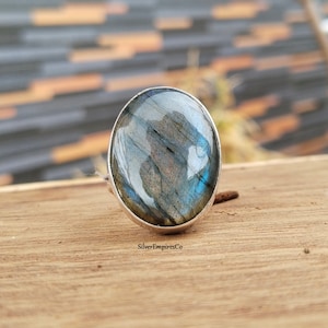 Könnte beinhalten: Ovaler Labradorit-Ring mit Silberband. Der Edelstein zeigt schillernde blaue, graue und braune Farbtöne. Der Ring liegt auf einer Holzoberfläche. Der Text "SilverEmpiresCo" ist sichtbar.