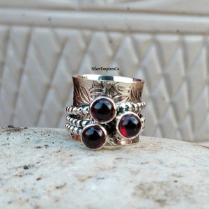 Könnte beinhalten: Ein silberfarbener Spinner-Ring mit drei dunkelroten Edelsteinen. Der Ring hat ein eingraviertes Blumenmuster auf dem oberen Band und Perlendetails auf den sich drehenden Bändern. Der Ring wird auf einer strukturierten Oberfläche gezeigt.