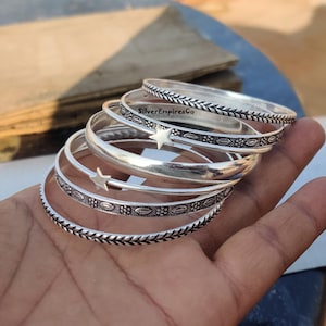 Pode incluir: Um conjunto de cinco pulseiras de prata com diferentes desenhos gravados. As pulseiras estão empilhadas umas sobre as outras e apresentam um desenho de estrela na pulseira superior.