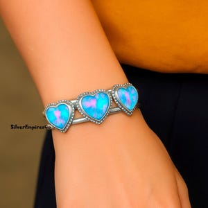 May include: A silver cuff bracelet featuring three heart-shaped opal stones with a blue and pink color scheme. Each heart is outlined with small, clear crystals. The bracelet is a stylish piece of jewelry.