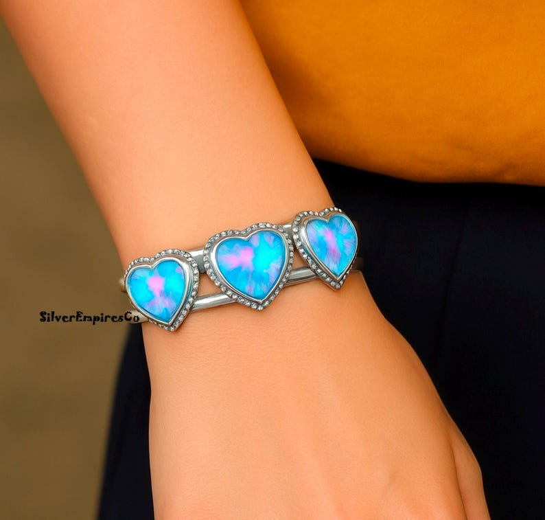 May include: A silver cuff bracelet featuring three heart-shaped opal stones with a blue and pink color scheme. Each heart is outlined with small, clear crystals. The bracelet is a stylish piece of jewelry.