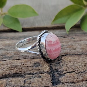 Könnte beinhalten: Ein silberner Ring mit einem ovalen, rosa und weiß gestreiften Edelstein. Der Ring hat ein seilartiges Design um die Band und der Text "SilverEmpiresCo" ist in die Band graviert.