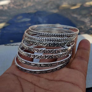 May include: A stack of silver bangles with intricate floral and leaf patterns. The bangles are held in a hand, showcasing the detailed designs and the shiny, reflective surface of the metal. The bangles vary in design, with some featuring braided textures.
