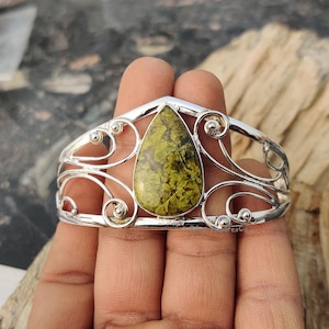 May include: A silver cuff bracelet with a green and white teardrop-shaped gemstone set in the center. The bracelet has a delicate, ornate design.